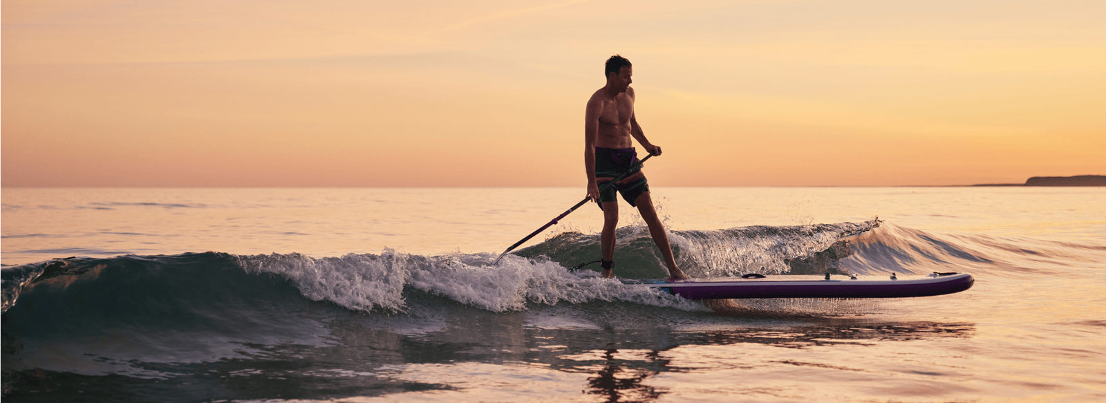 A man paddleboarding, standing up in the sea at sunset