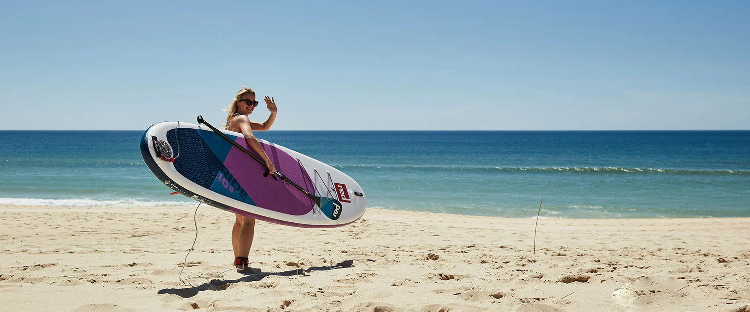 A woman on the beach carrying her paddleboard, waving at someone