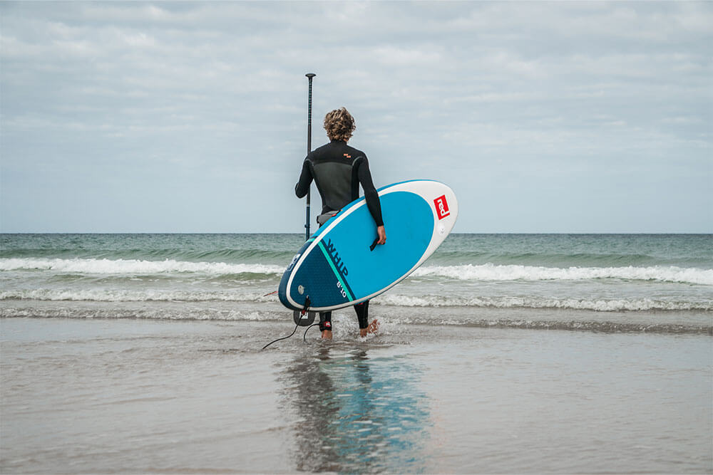 man wearing wetsuit walking into the sea carrying Whip MSL inflatable paddle board