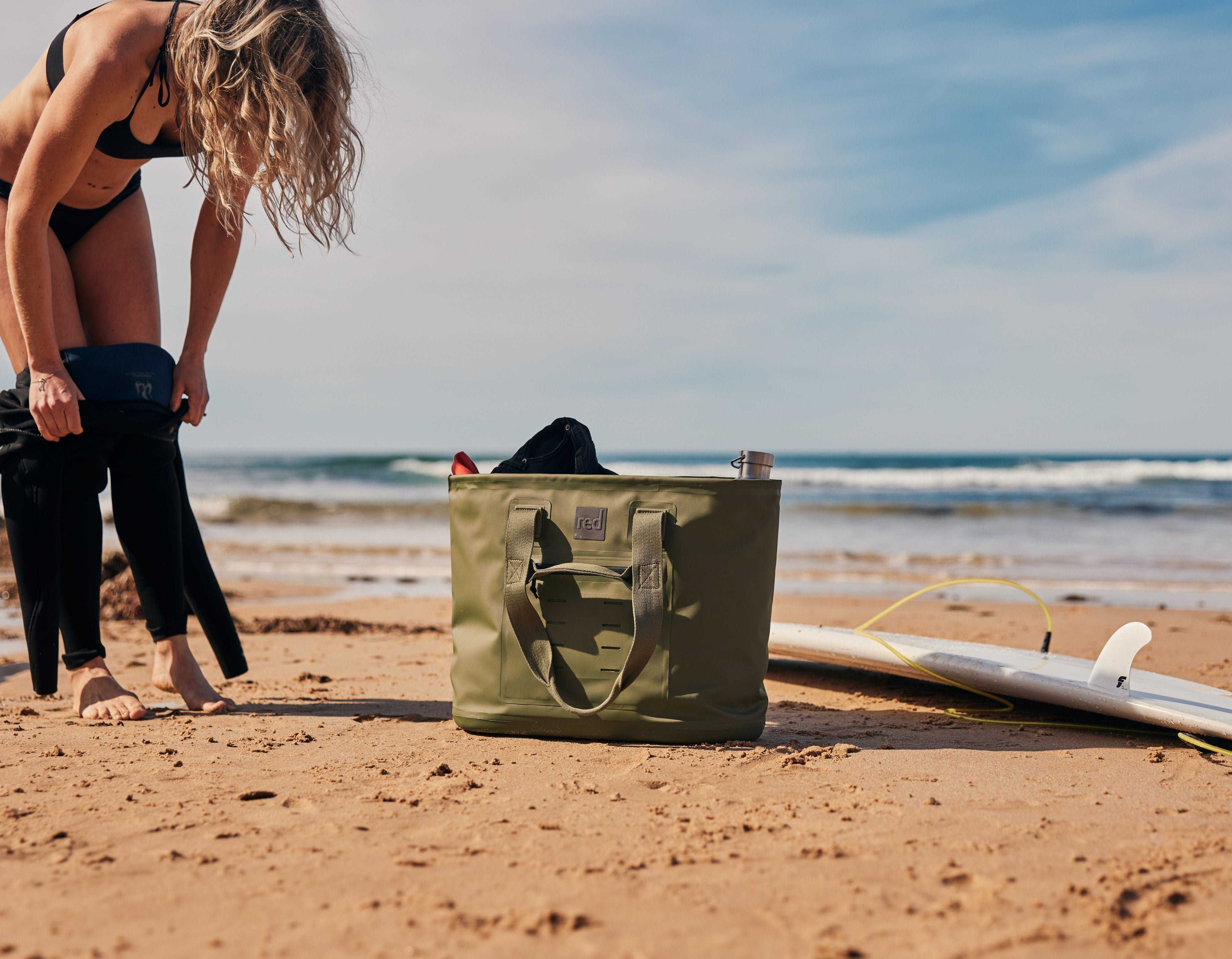 Woman changing on the beach beside a surf board and a Red Olive Green Waterproof Tote Bag. 